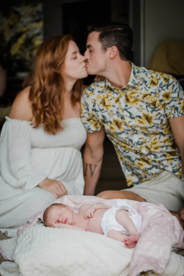 New parents kissing on the couch with their baby asleep on a pillow in front of them