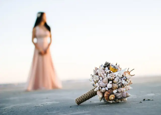 Seashell bouquet resting on the sand in focus, a mother-to-be in a blush gown blurred behind it