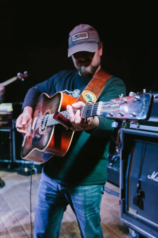 Singer playing acoustic guitar on stage in a ball cap