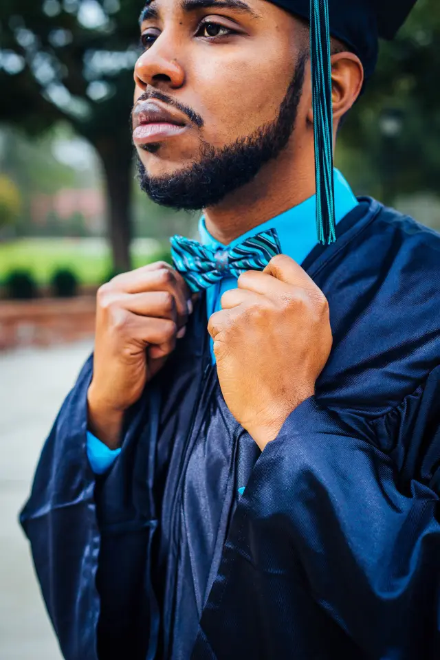 Graduate in a navy gown and teal bow tie adjusting his collar, looking off to the side