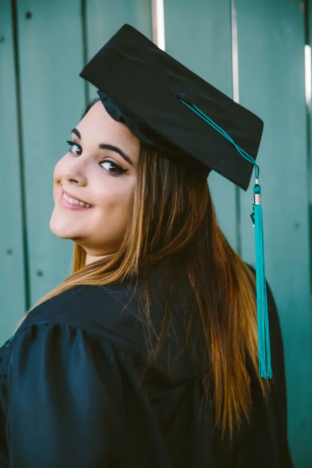 Graduate in a cap with a teal tassel smiling over her shoulder by a teal fence