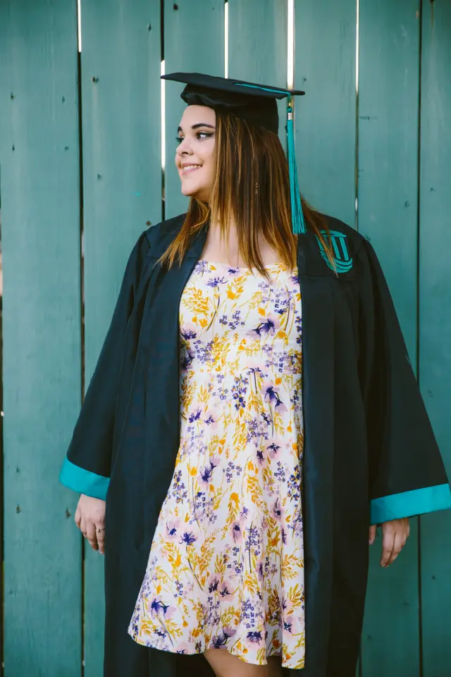 Graduate in her open gown over a floral dress beside a teal fence