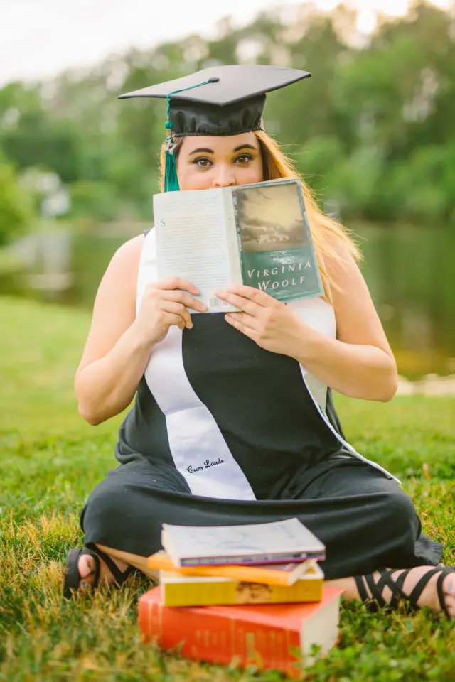 Graduate sitting in the grass reading a book with a stack of books beside her