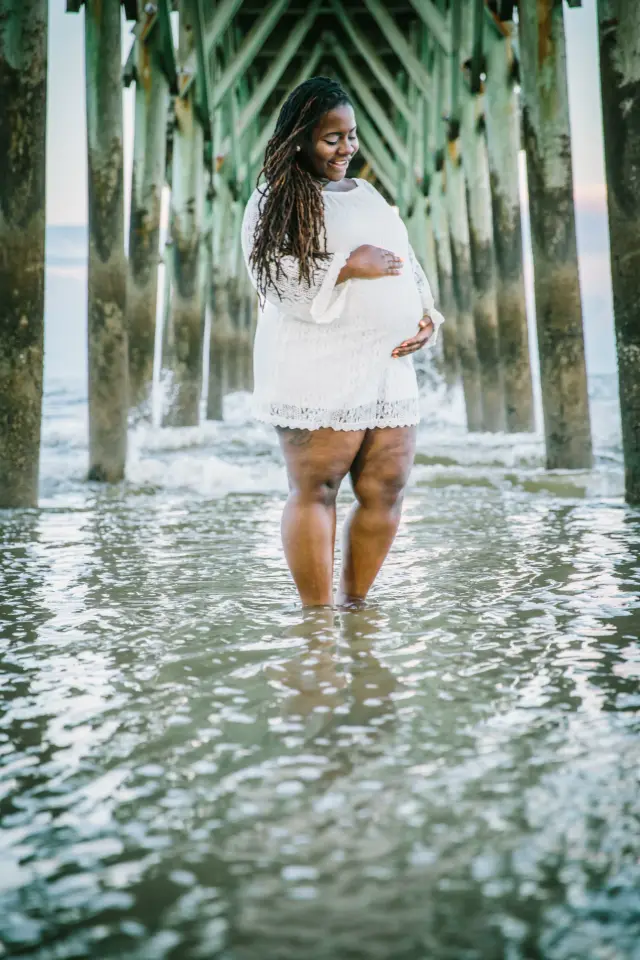 Mother-to-be in a white lace dress standing in the shallow water under the pier