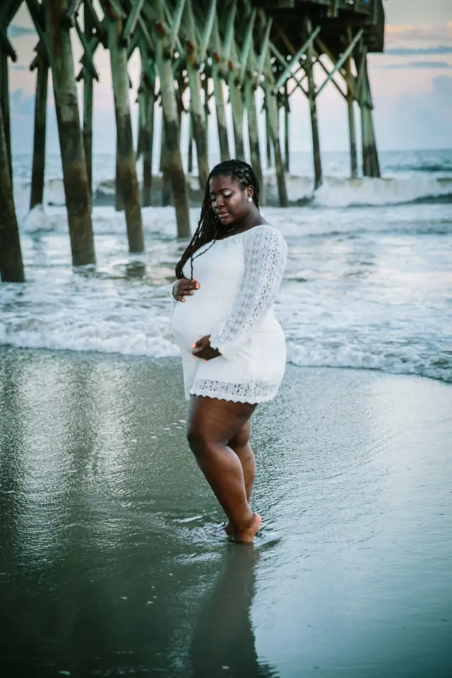 Mother-to-be in white lace walking through the surf by the pier pilings