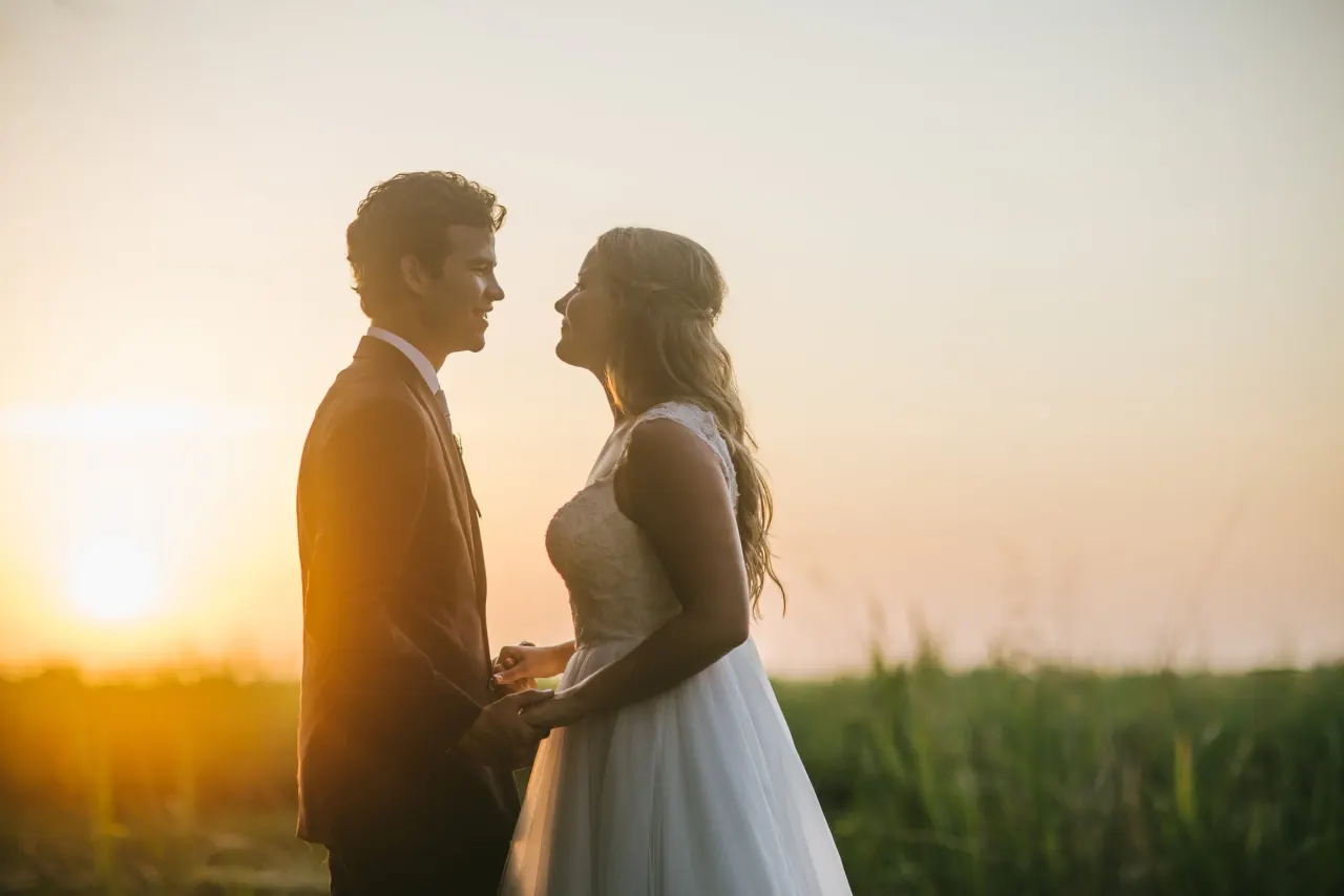 Bride and groom facing each other in silhouette at sunset in the dune grass