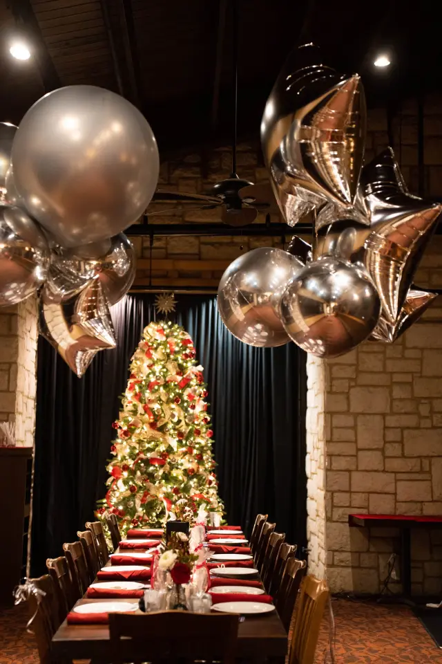 Holiday party long table set beside a Christmas tree under silver and gold balloons