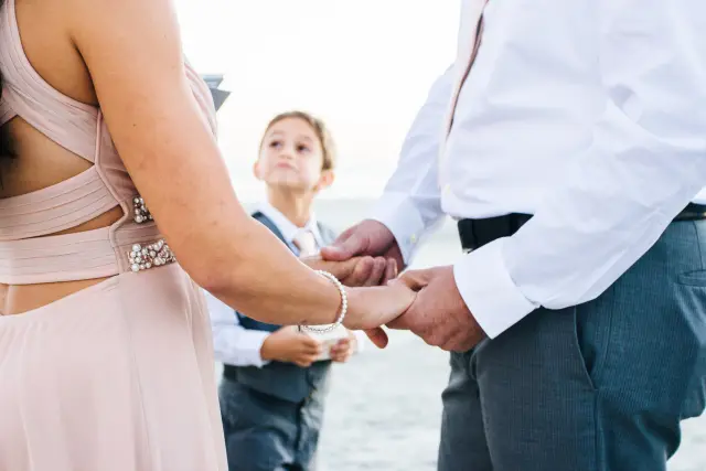 Close-up of the couple holding hands during their beach vows as a young ring bearer looks on