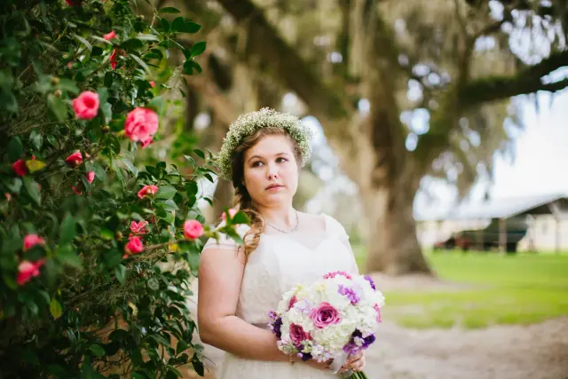 Bride with a flower crown holding a purple and white bouquet beside camellias and live oaks