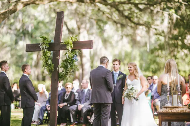 Outdoor wedding ceremony under live oaks and Spanish moss, with a wooden cross at the altar