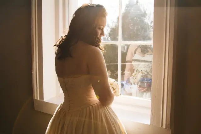 Bride in silhouette by a sunlit window, looking down at her bouquet