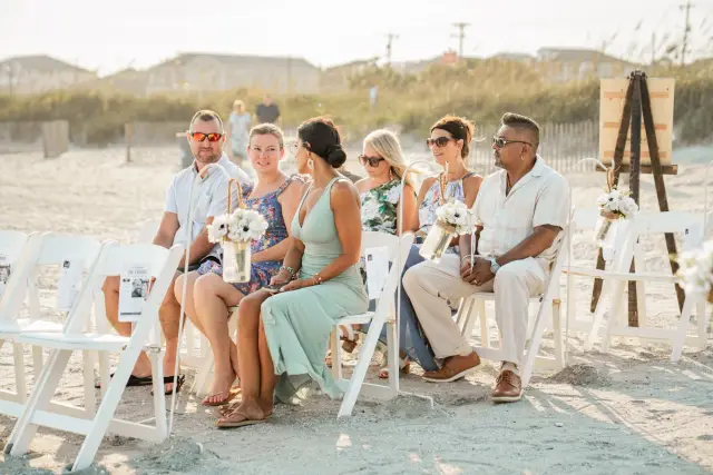 Guests seated in white chairs on the sand waiting for a beach ceremony to begin