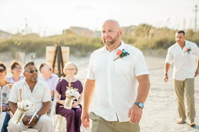 Groom in a white shirt walking up the sandy aisle to his beach ceremony
