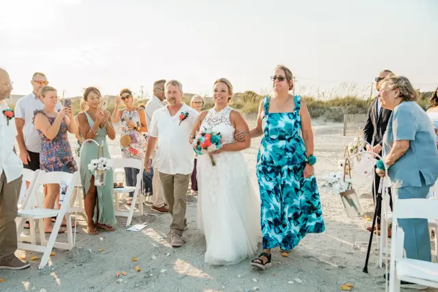 Bride walking down the sandy aisle with her mother as guests stand