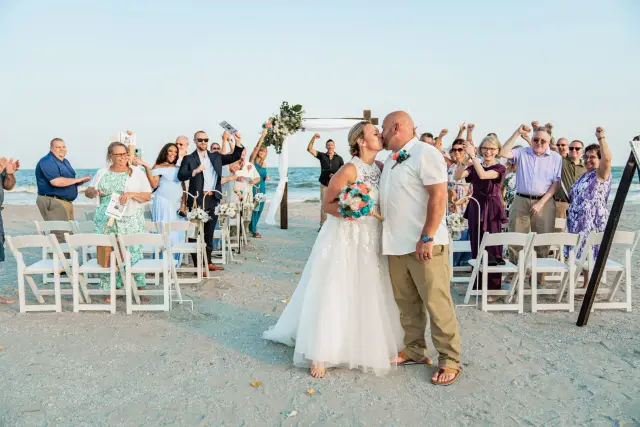 Bride and groom kissing at the end of the aisle as guests cheer on the beach