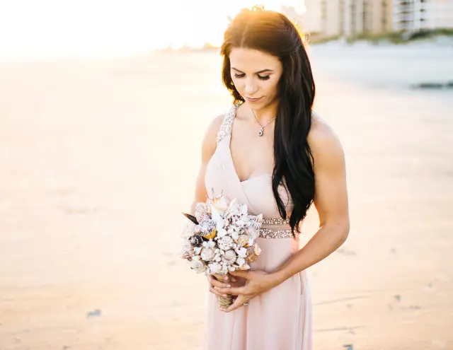 Bride in a blush gown looking down at her seashell bouquet on the beach in morning light