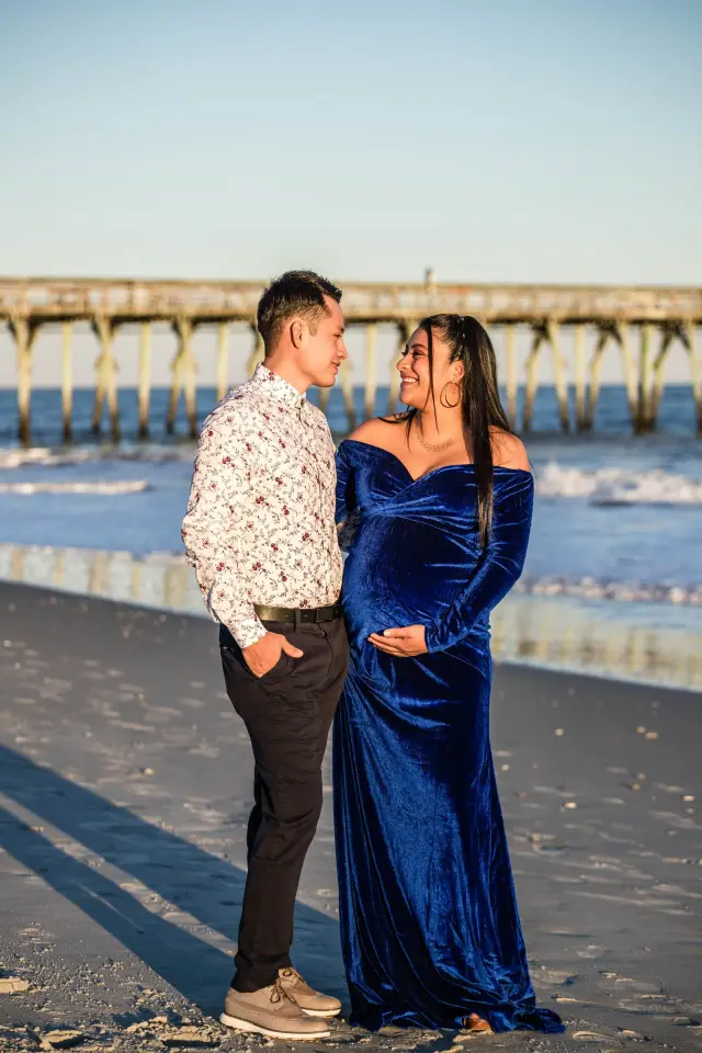 Expecting couple holding hands by the fishing pier, her blue gown trailing on the sand