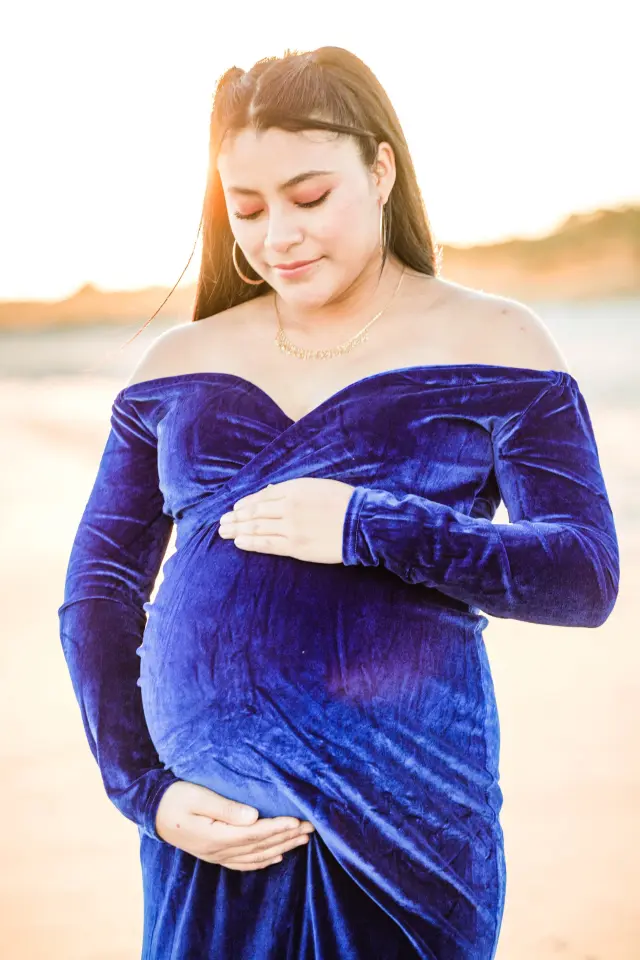Mother-to-be in a blue velvet off-shoulder gown cradling her belly, backlit by the sun