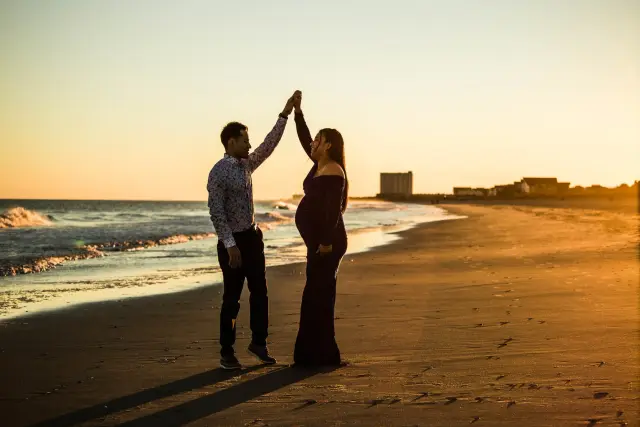 Expecting couple dancing on the beach at sunset, silhouetted against a golden sky