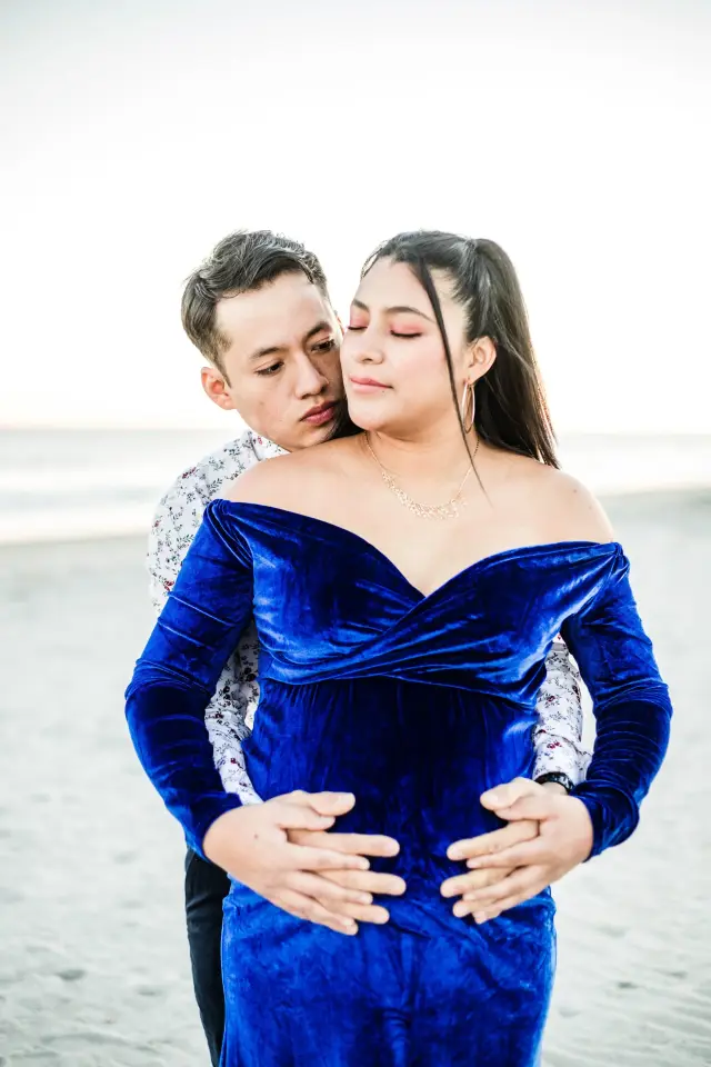 Partner holding the mother-to-be from behind, both hands on her belly, on the beach