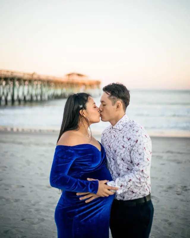 Expecting couple kissing on the beach with the pier behind them at dusk