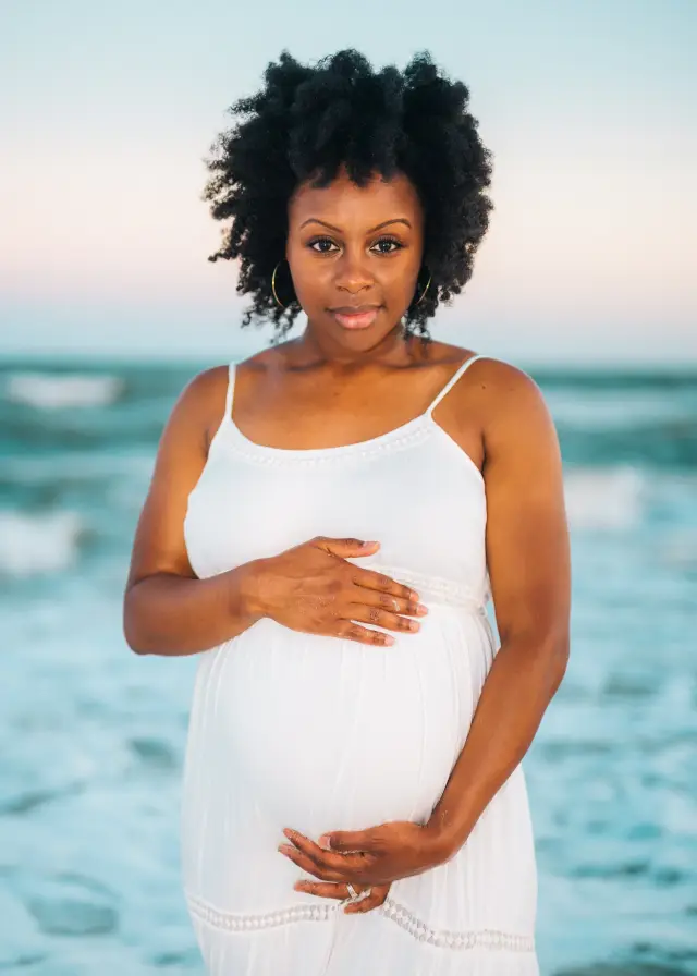Mother-to-be in a white sundress cradling her belly with the ocean behind her
