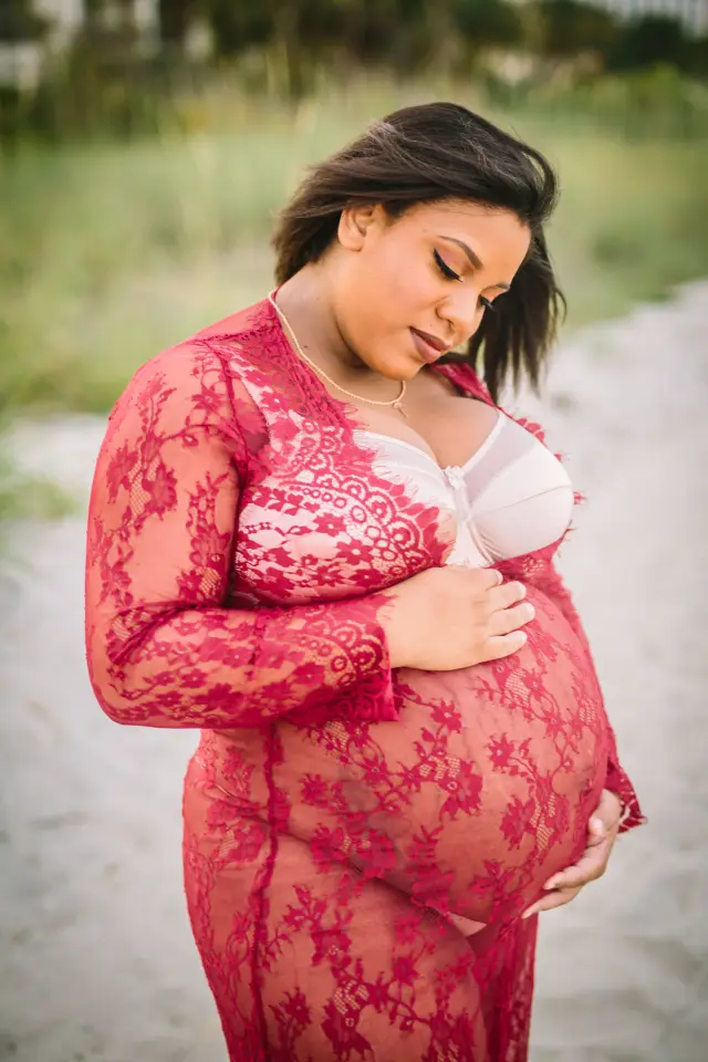 Mother-to-be in a red lace robe looking down at her belly in the dunes