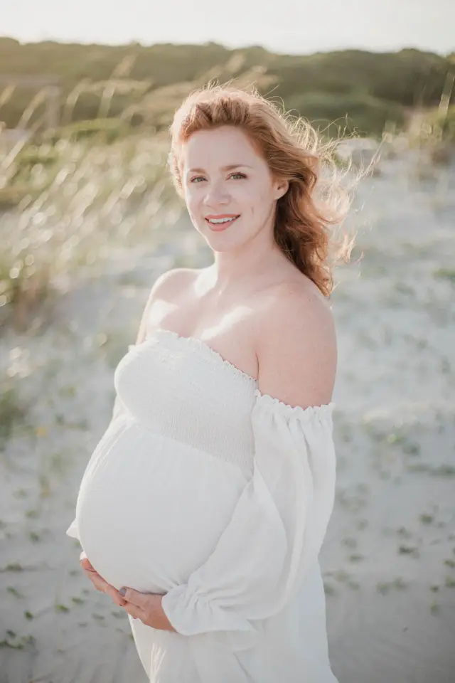 Red-haired mother-to-be in a white off-shoulder dress smiling in the dune grass