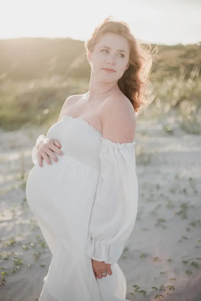 Mother-to-be in a white dress looking over her shoulder in the sea oats