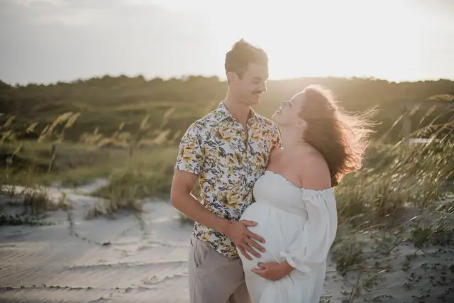 Expecting couple laughing together in the dunes, his hand on her belly