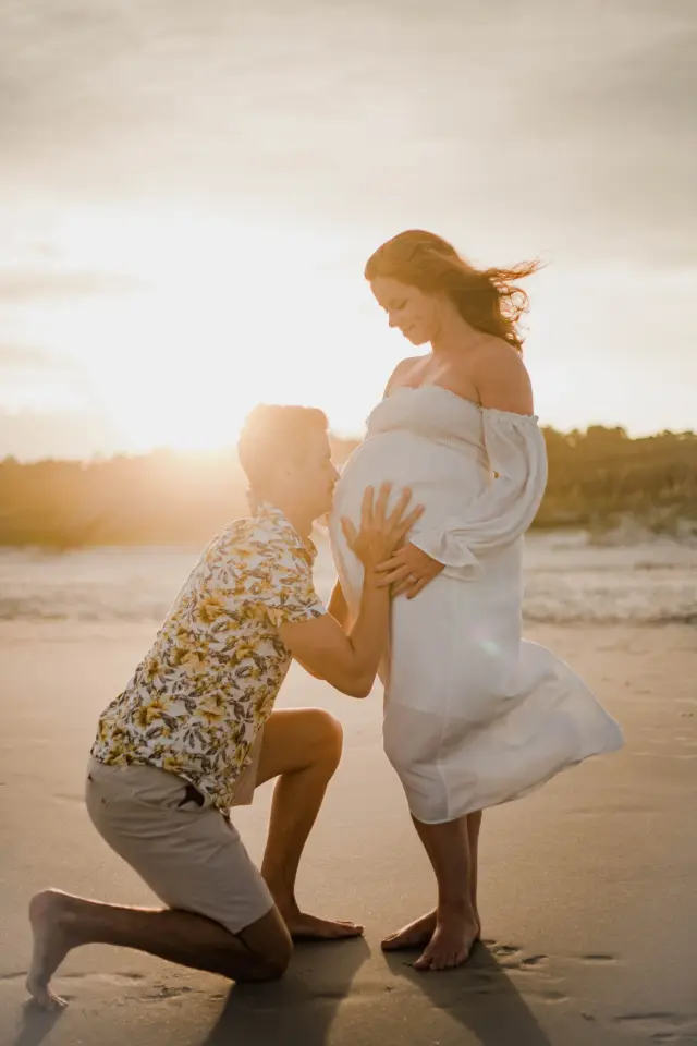 Dad-to-be kneeling to kiss the baby bump on the beach at sunset