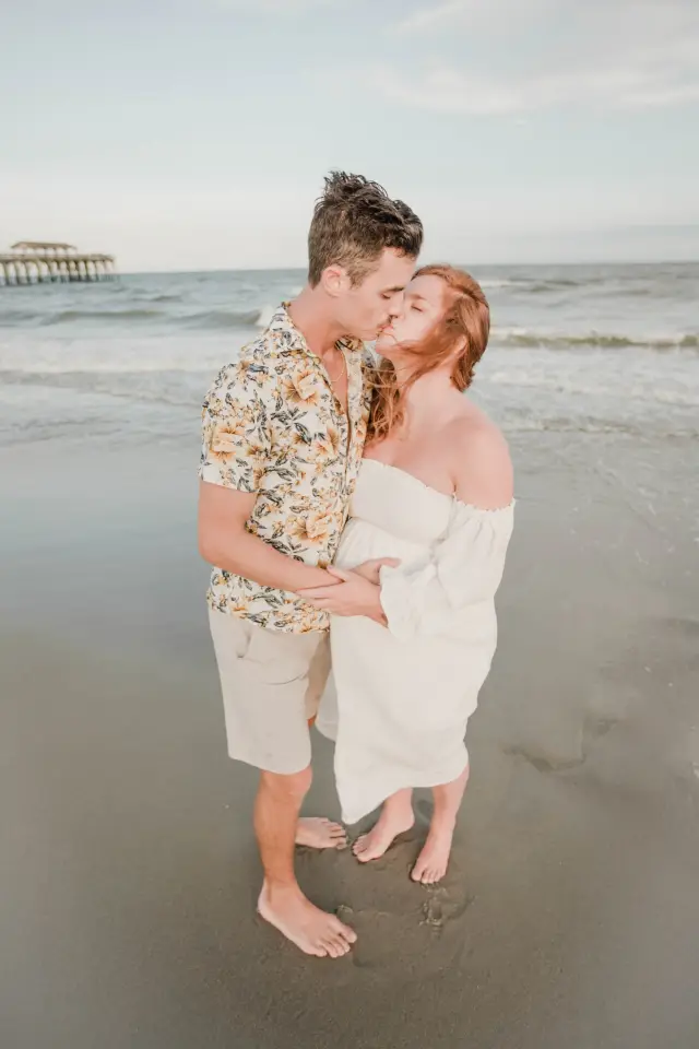 Expecting couple kissing at the water's edge with the pier in the distance