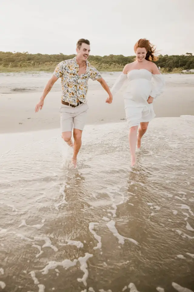 Expecting couple running hand in hand through the shallow surf